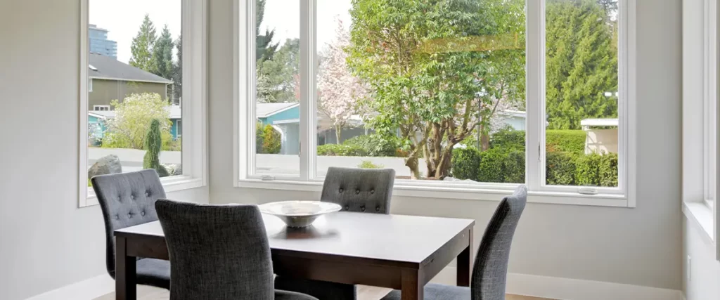 A sleek dining space with modern lighting, gray chairs, and expansive windows offering a view of lush greenery.