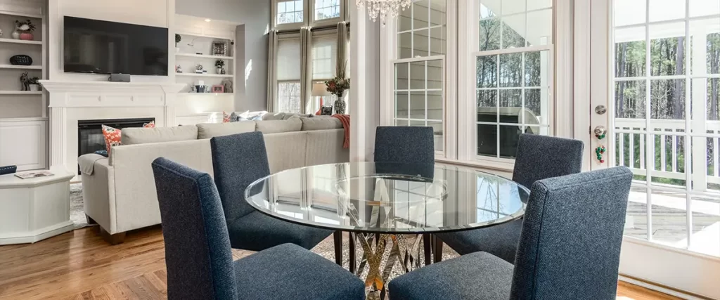 A stylish dining space with glass table, navy chairs, and large white-trimmed windows leading to a bright outdoor deck.