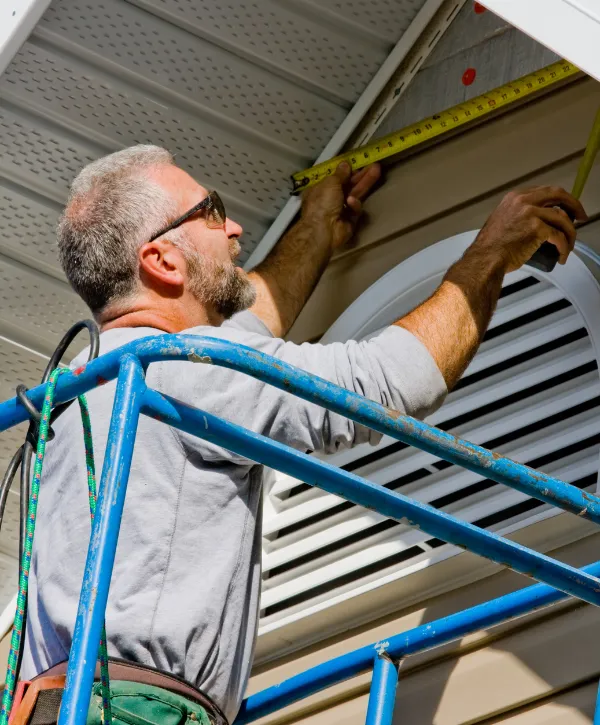 A contractor on scaffolding using a measuring tape to install soffit and siding on a house.