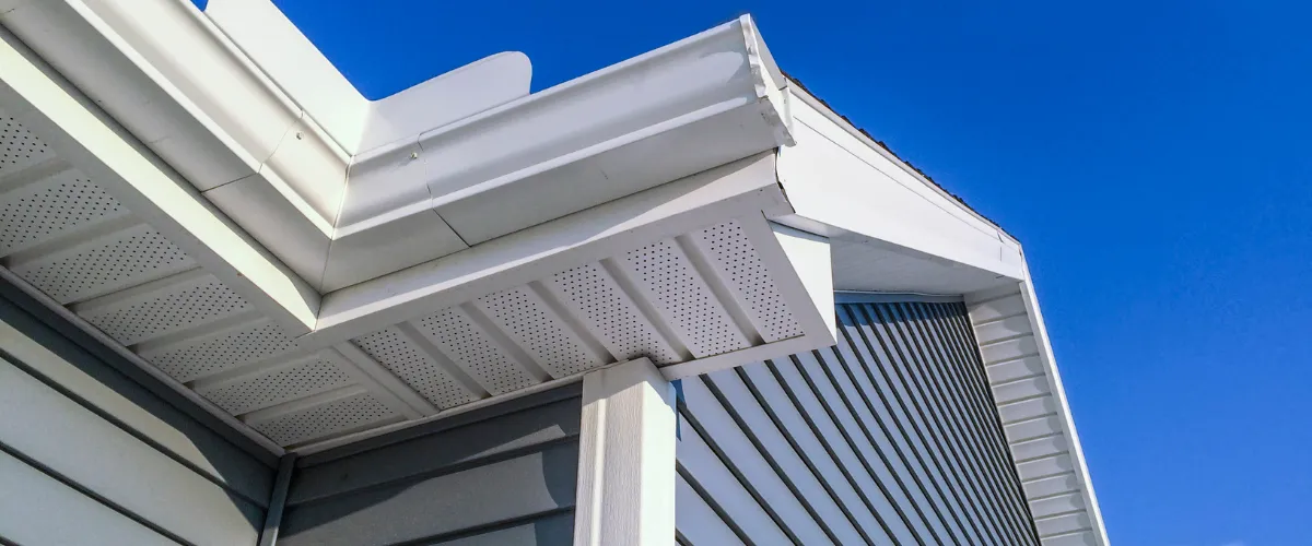 "A house exterior showing white soffit, fascia, and gutters set against blue-gray vinyl siding under a clear blue sky.