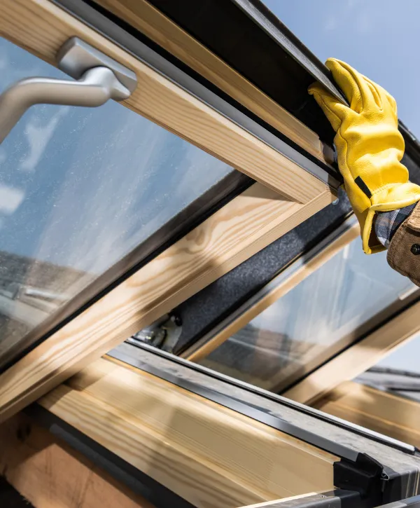 A close-up of a worker wearing yellow gloves installing a roof window with a wooden frame