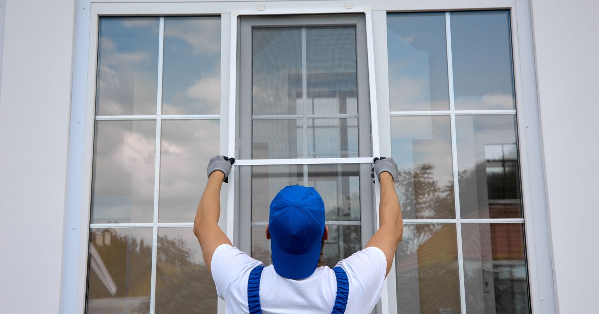 A worker in a blue cap and overalls installing a window screen on a large glass window.