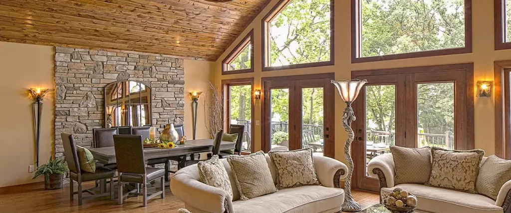 Rustic living room with vaulted wood ceiling, stone fireplace, and large picture windows overlooking wooded backyard