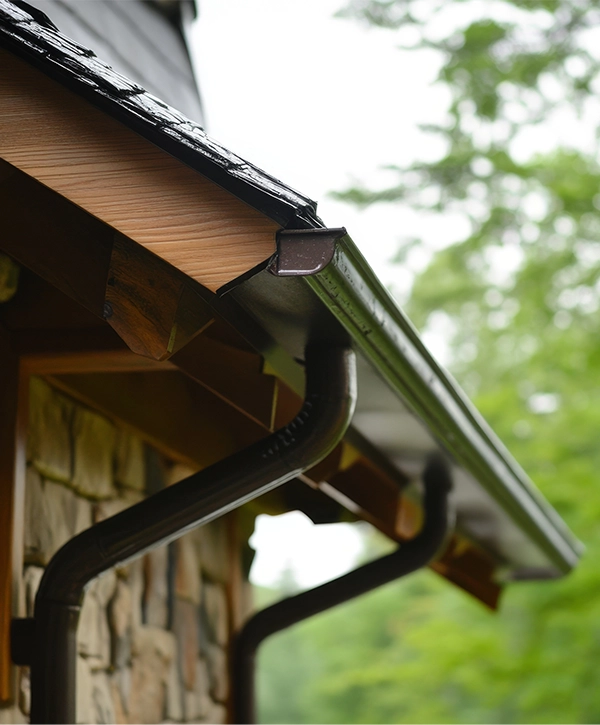 Close-up of a perfectly installed brown seamless gutter and downspout on the corner of a house with stone and wood-shake siding.