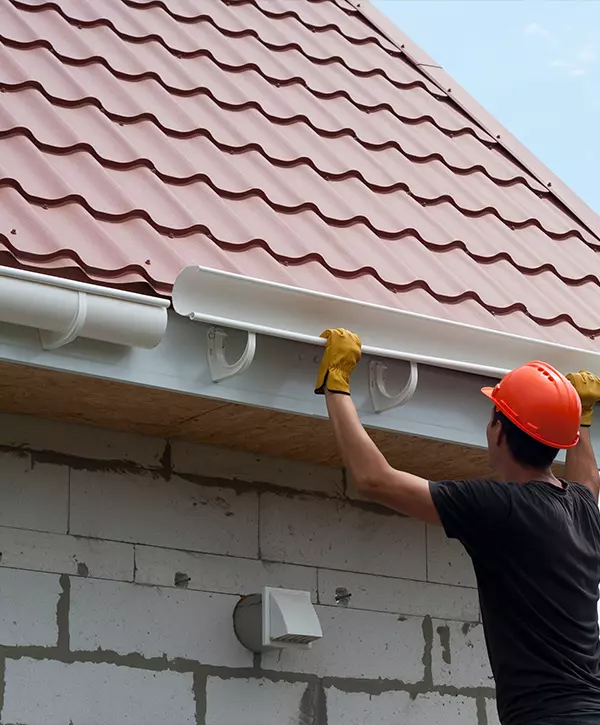 Construction worker wearing an orange safety helmet installing a section of white half-round gutter beneath a red metal tiled roof and a white brick wall.