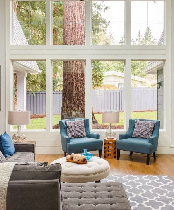Spacious living room with high ceilings and tall multi-pane windows providing a view of mature trees, furnished with blue armchairs.