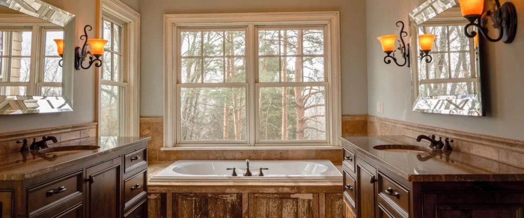 A spacious, luxury bathroom featuring a white soaking tub centered beneath a large double-hung window looking out onto winter trees, flanked by dark wood vanity cabinets.