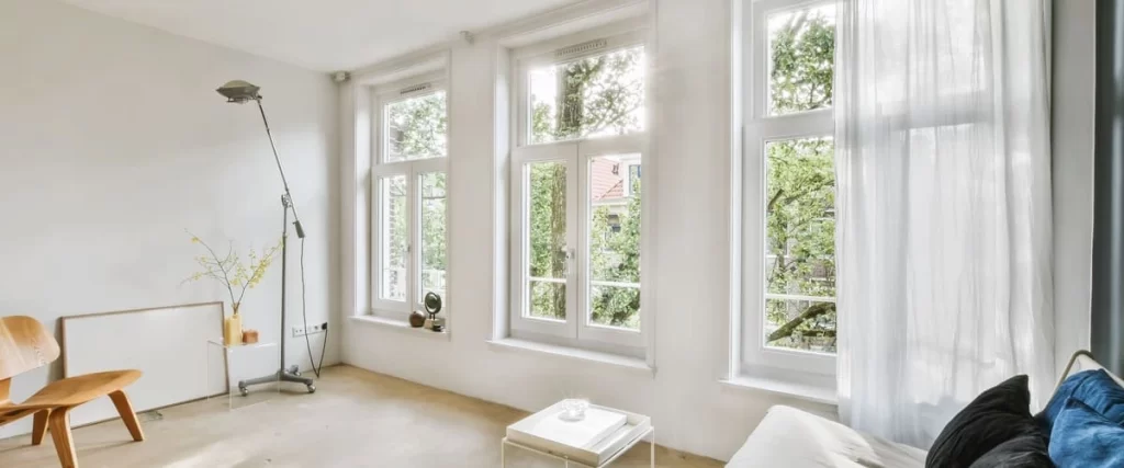 Interior view of three tall white double-hung windows in a minimalist living space overlooking lush green trees.