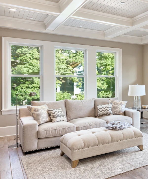 A bright, comfortable living room featuring a beige sofa and ottoman, showcasing a recent window replacement in Lincoln, NE with three large vertical windows looking out at green trees.