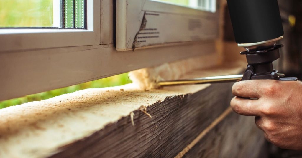 A worker using a spray gun applicator to apply expanding polyurethane foam around windows to seal gaps on a wooden sill.